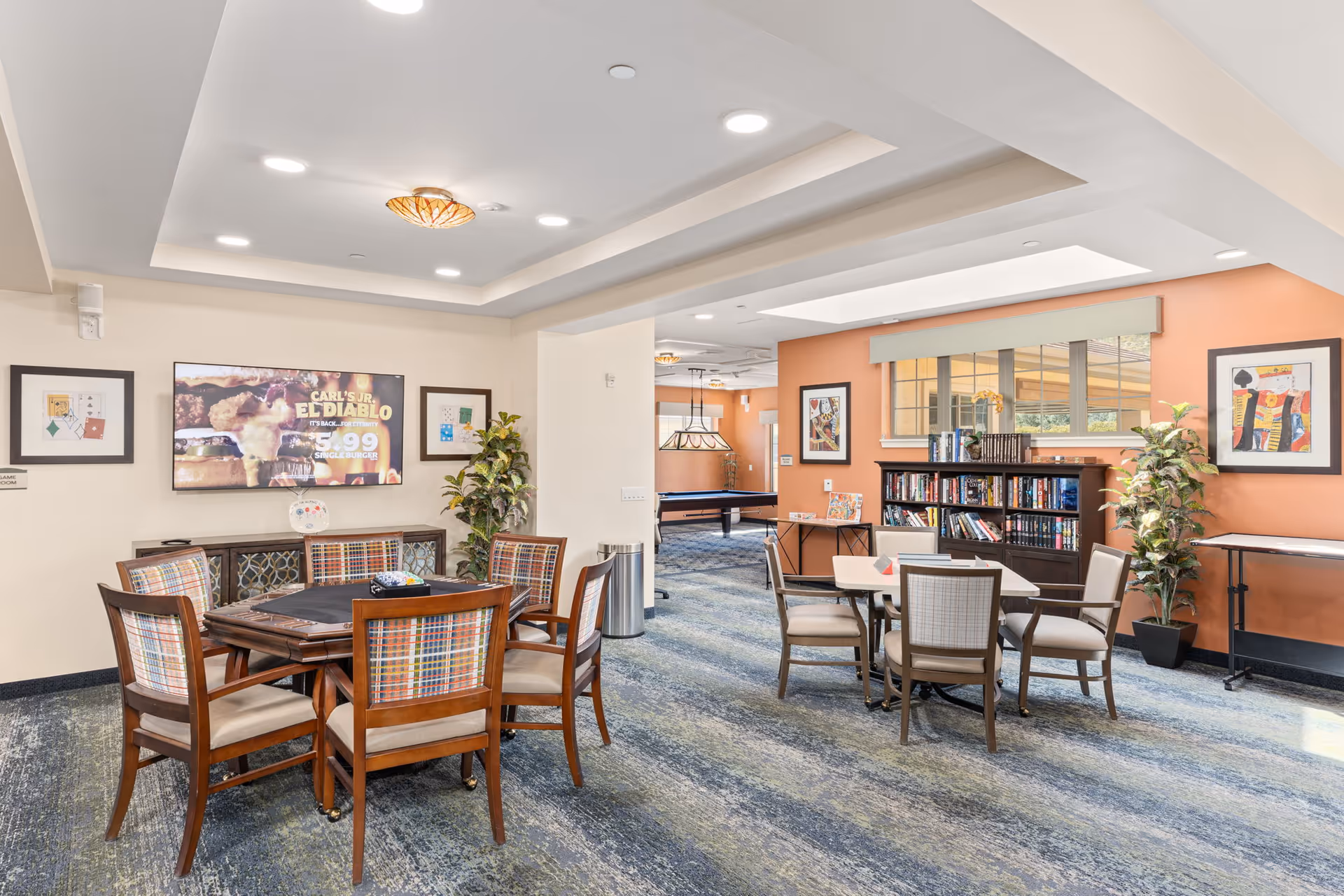 Bright communal activity room with card tables and chairs, bookshelves, a wall-mounted TV, and a pool table in the background.