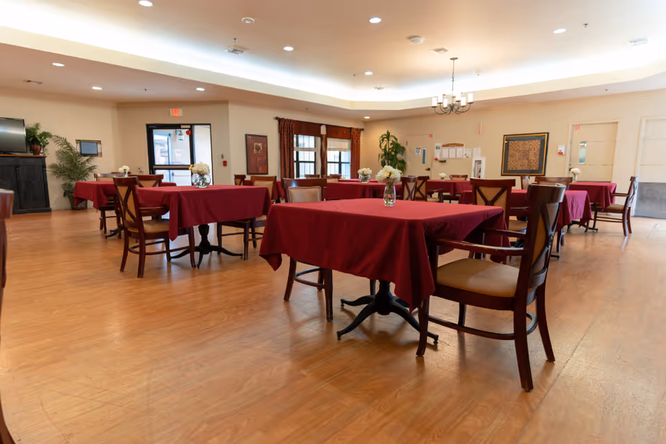 A spacious dining room with multiple tables covered in red tablecloths, each table surrounded by wooden chairs with cushioned seats. Small flower arrangements are placed on each table. The room has wooden flooring, beige walls, and a ceiling with recessed lighting and a chandelier. There are windows with curtains and a door visible in the background.