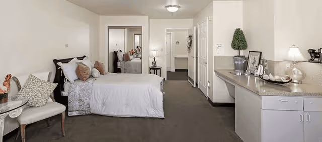 Spacious senior living studio room with a bed, seating, kitchenette counter, mirrored closet, and hallway.