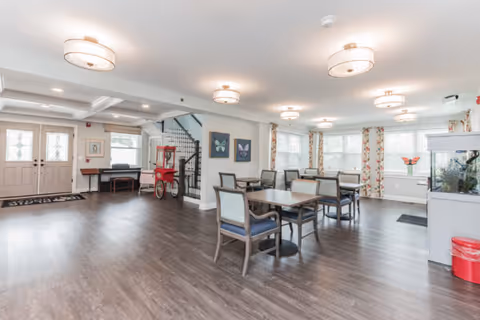 Spacious bright communal dining and activity room with several tables and chairs, a red popcorn cart by the entrance, and large windows with floral curtains.