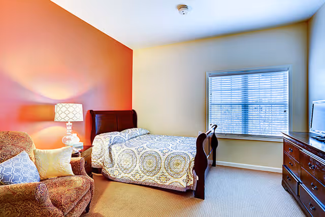 A cozy bedroom with a single bed featuring a patterned bedspread, a wooden headboard and footboard, a wooden dresser with a TV on top, a window with blinds, a patterned armchair with pillows, and a side table with a lamp. One wall is painted orange while the others are light beige.