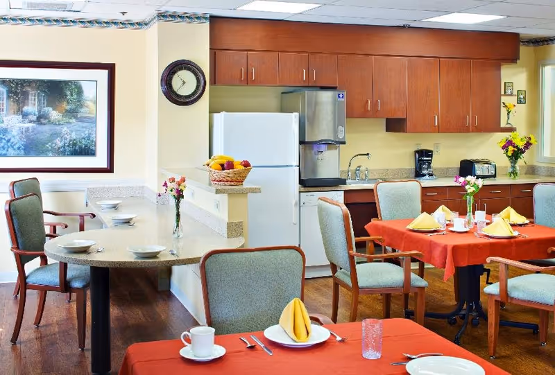 A bright and clean dining area in a senior living facility with tables covered in red tablecloths, set with plates, cups, utensils, and yellow folded napkins. There are several chairs around the tables. In the background, there is a kitchen area with wooden cabinets, a white refrigerator, a dishwasher, a coffee maker, a toaster, and a basket of fruit on the counter. A wall clock and a framed painting are visible on the wall.