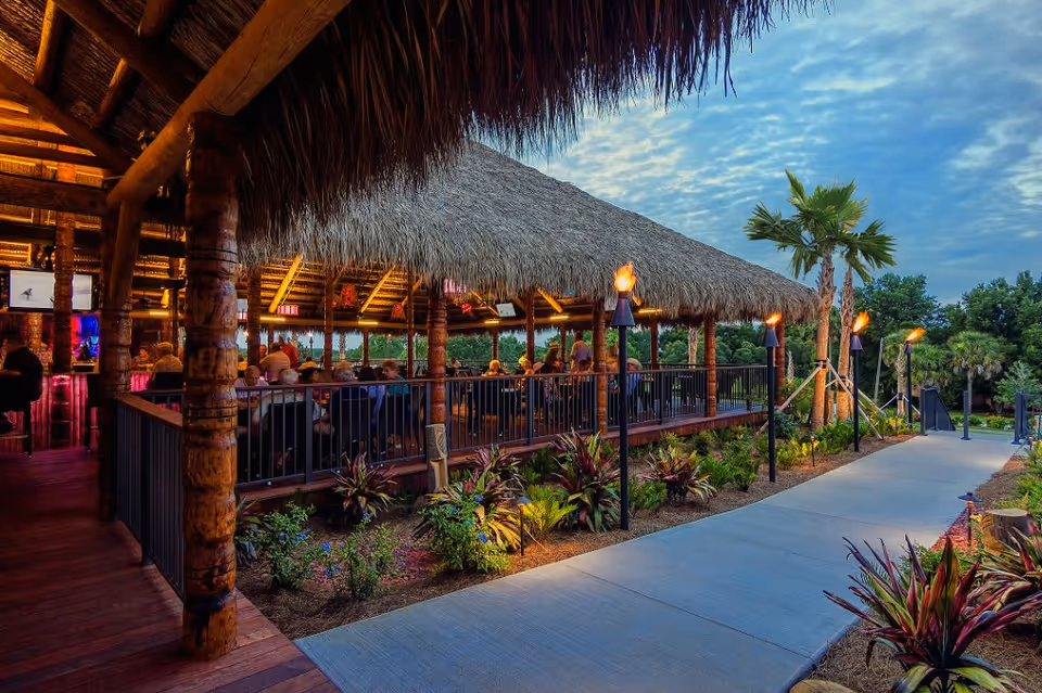 Thatched-roof outdoor tiki pavilion with people dining under warm lights beside a palm-lined walkway.