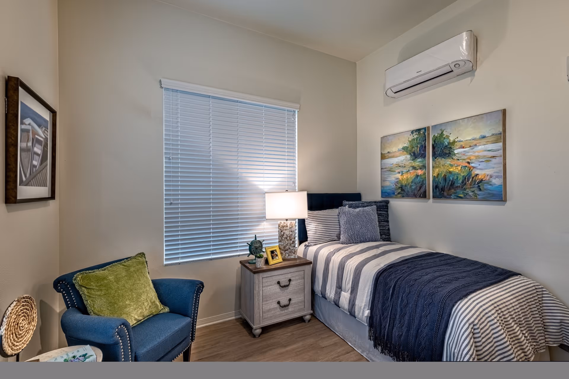 A cozy bedroom with a single bed featuring striped bedding and multiple pillows. Next to the bed is a wooden nightstand with a lamp, a small plant, and a framed photo. A blue armchair with a green cushion is positioned near the nightstand. The room has a window with closed blinds, two landscape paintings on the wall above the bed, and an air conditioning unit mounted on the wall.