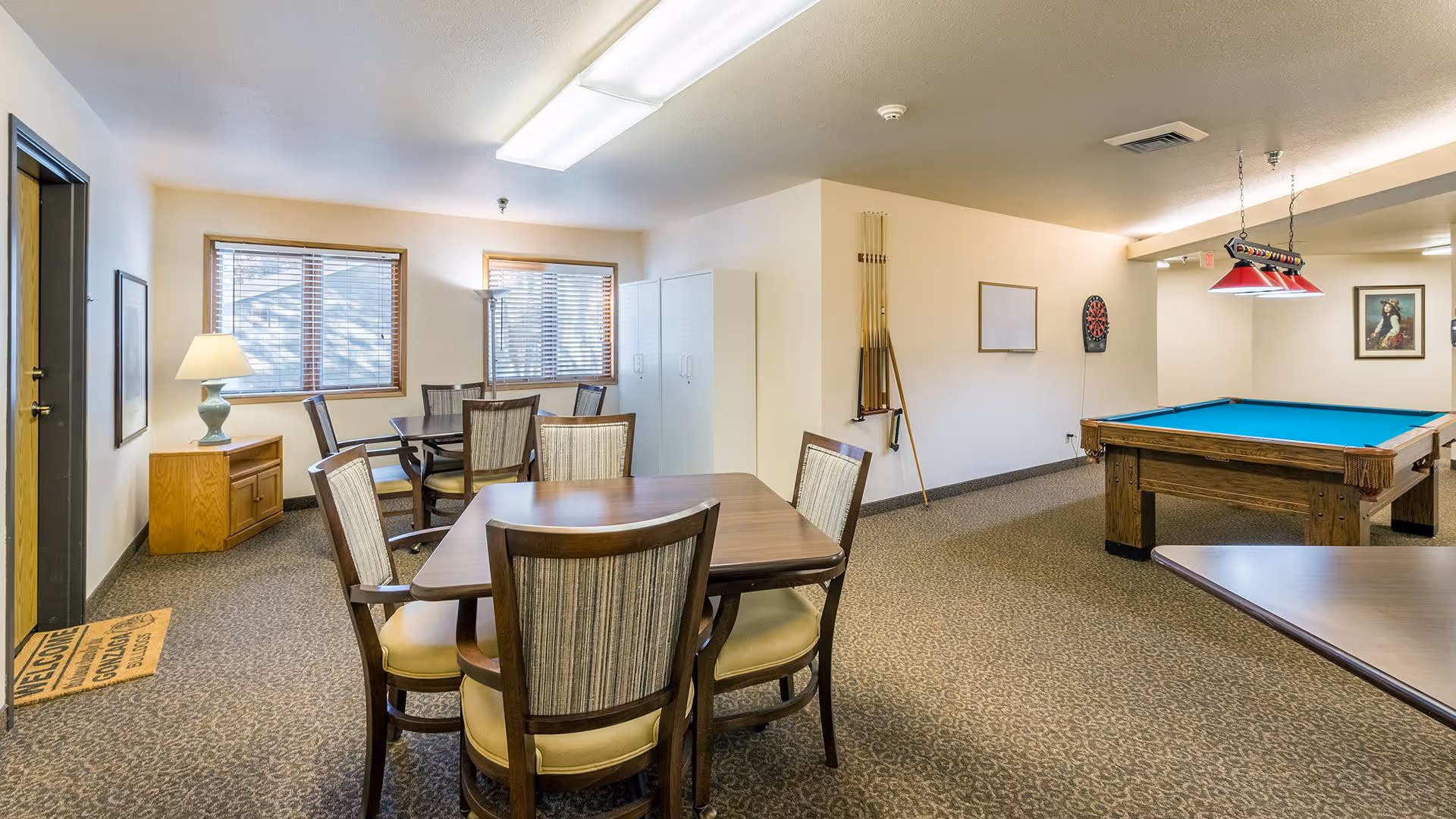 A spacious common room with multiple tables and chairs, a pool table with overhead lighting, a dartboard on the wall, and windows letting in natural light. The room has carpeted floors, neutral-colored walls, and a lamp on a small wooden cabinet near the entrance door.