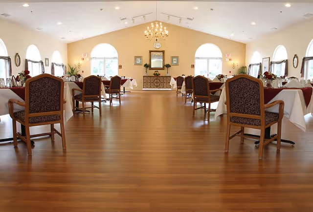 A spacious dining room with wooden floors, several tables covered with white and burgundy tablecloths, and chairs with patterned upholstery. The room has large arched windows letting in natural light, a chandelier hanging from the ceiling, and decorative plants and framed pictures on the walls.