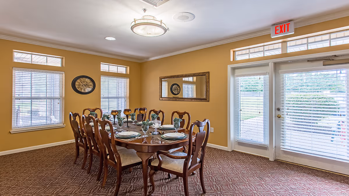 A dining room with a wooden dining table set for eight people. The table is set with plates, green and amber glasses, and napkins. The room has yellow walls, two windows with white blinds, a large rectangular mirror on one wall, a round wall clock, and a glass door with an exit sign above it leading outside.