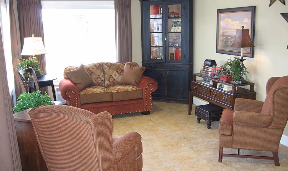 A cozy living room with a patterned loveseat, two upholstered armchairs, a wooden side table with a lamp and plant, a dark wooden corner cabinet with glass doors, and a wooden console table with books, a lamp, and a plant. The room has tiled flooring and light-colored walls with a framed painting.