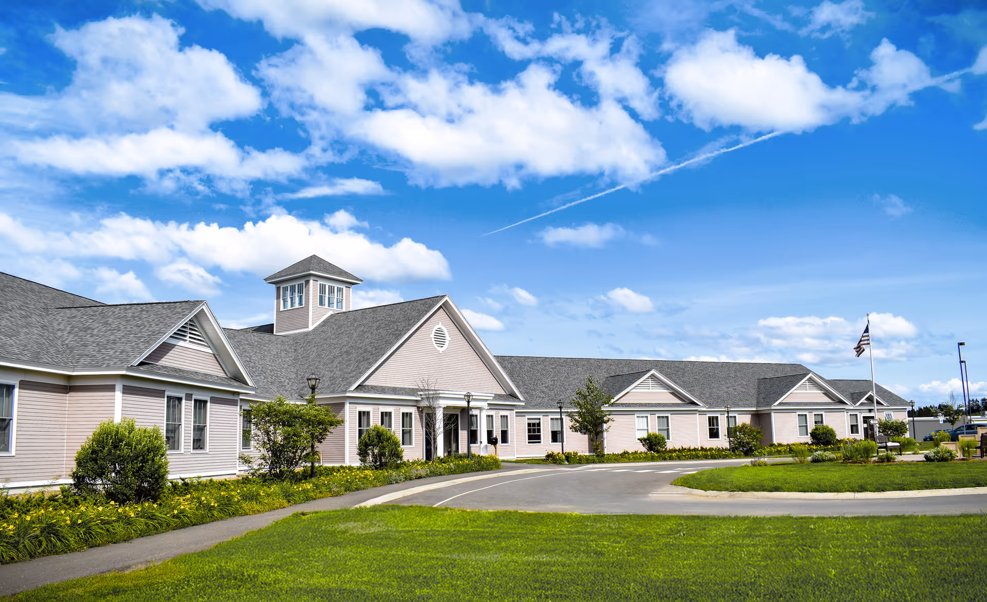 Low single-story beige senior living building with a circular driveway, landscaped lawn and an American flag under a blue sky.