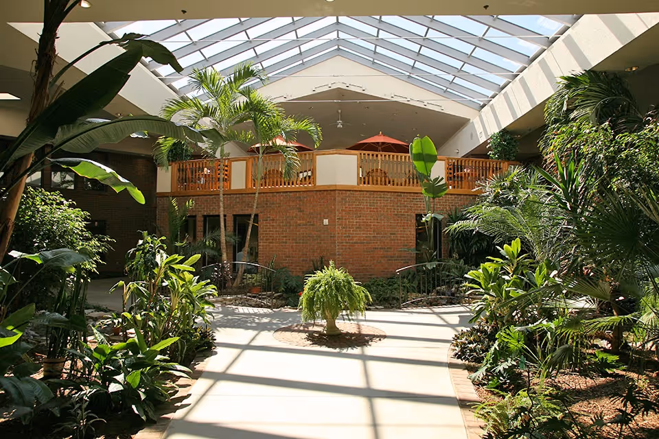 Indoor garden area with various green plants and palm trees under a large skylight ceiling. A brick structure with a wooden railing and red umbrellas is visible in the background.