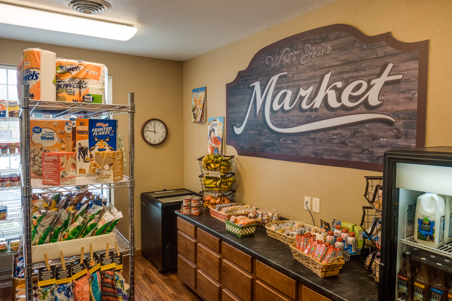 Interior view of a small market area labeled 'West Side Market' with shelves stocked with various grocery items including paper towels, cereals, snacks, bananas, and beverages. There is a clock on the wall and a refrigerator with milk and drinks.