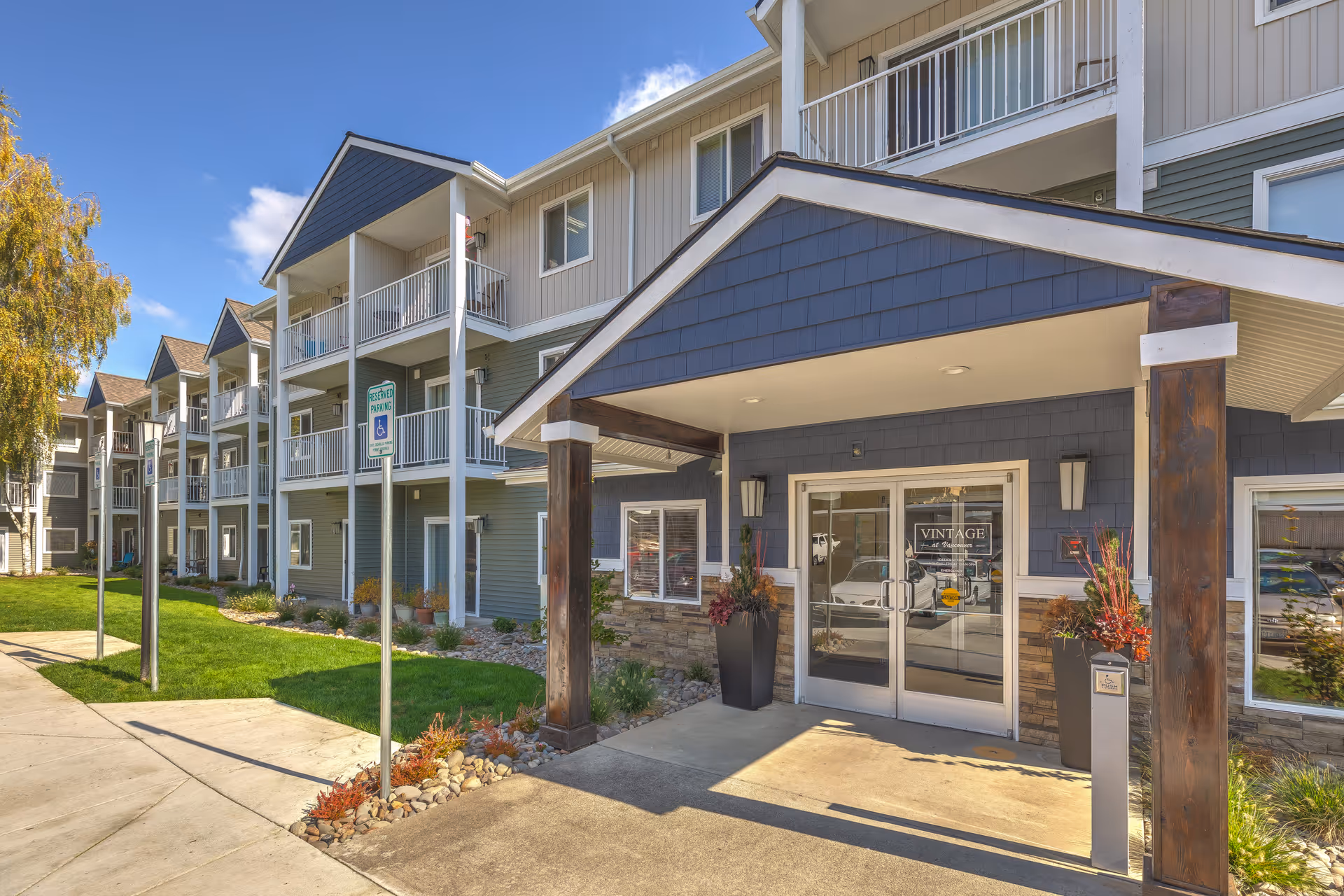 Exterior view of a multi-story senior living facility named Vintage at Vancouver, featuring a covered entrance with glass double doors, potted plants, and a well-maintained lawn with reserved parking signs.