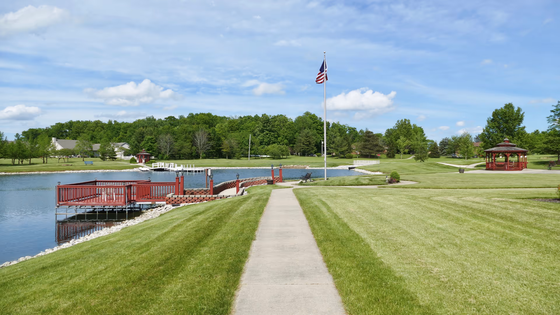 Well-manicured lawn and concrete path leading to a lakeside pier, flagpole, and gazebo under a blue sky.