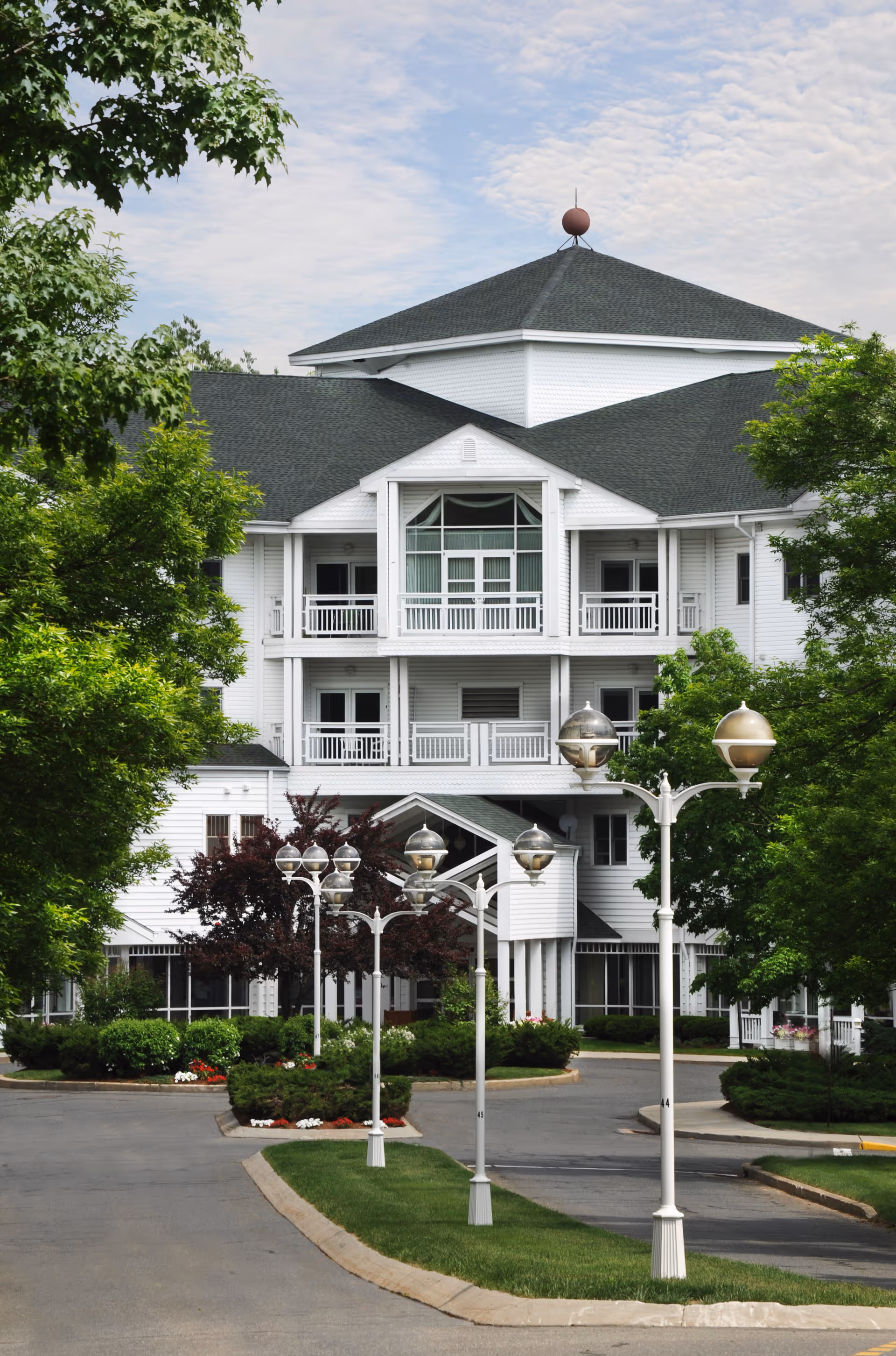 White multi-story senior living building with balconies, a landscaped driveway and decorative lamp posts framed by trees.