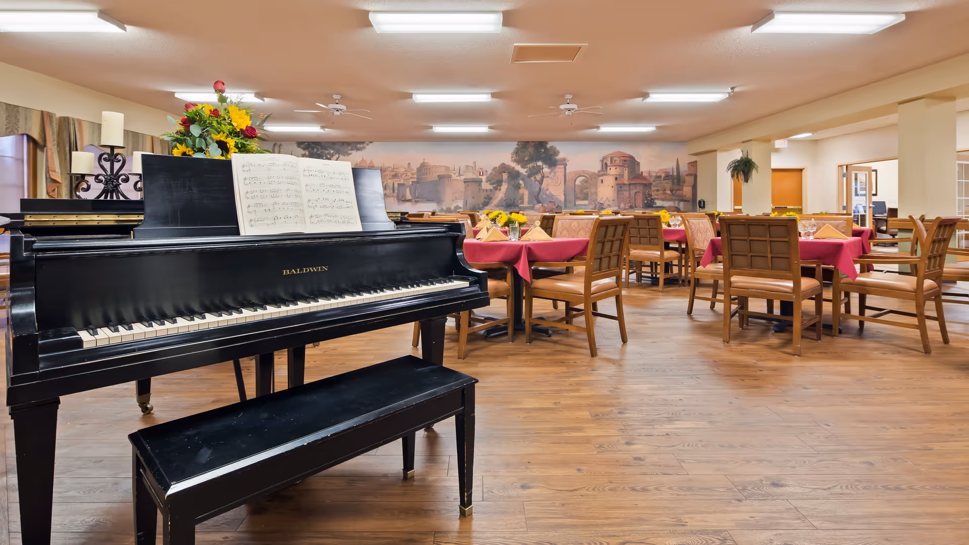 Interior view of a dining room with wooden floors, multiple tables covered with red tablecloths and set with napkins and glassware. A black Baldwin grand piano with sheet music and a floral arrangement is prominently placed in the foreground. The back wall features a mural depicting an ancient cityscape.