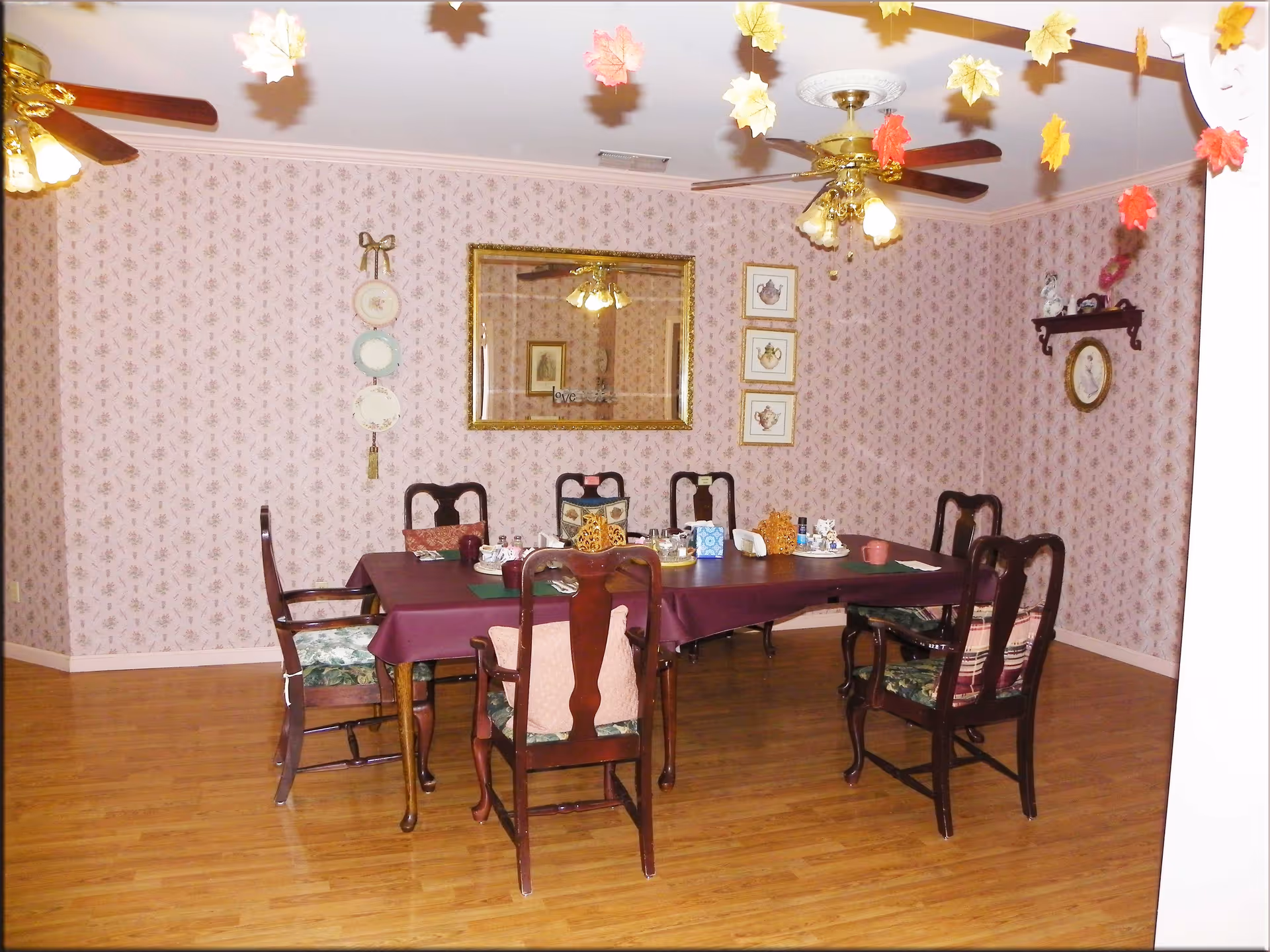 A dining room with a rectangular table covered with a purple tablecloth surrounded by six wooden chairs with cushions. The walls have floral wallpaper and are decorated with framed pictures, decorative plates, and a mirror. Two ceiling fans with lights hang from the ceiling, and autumn leaf decorations are suspended above the table. The floor is wooden.