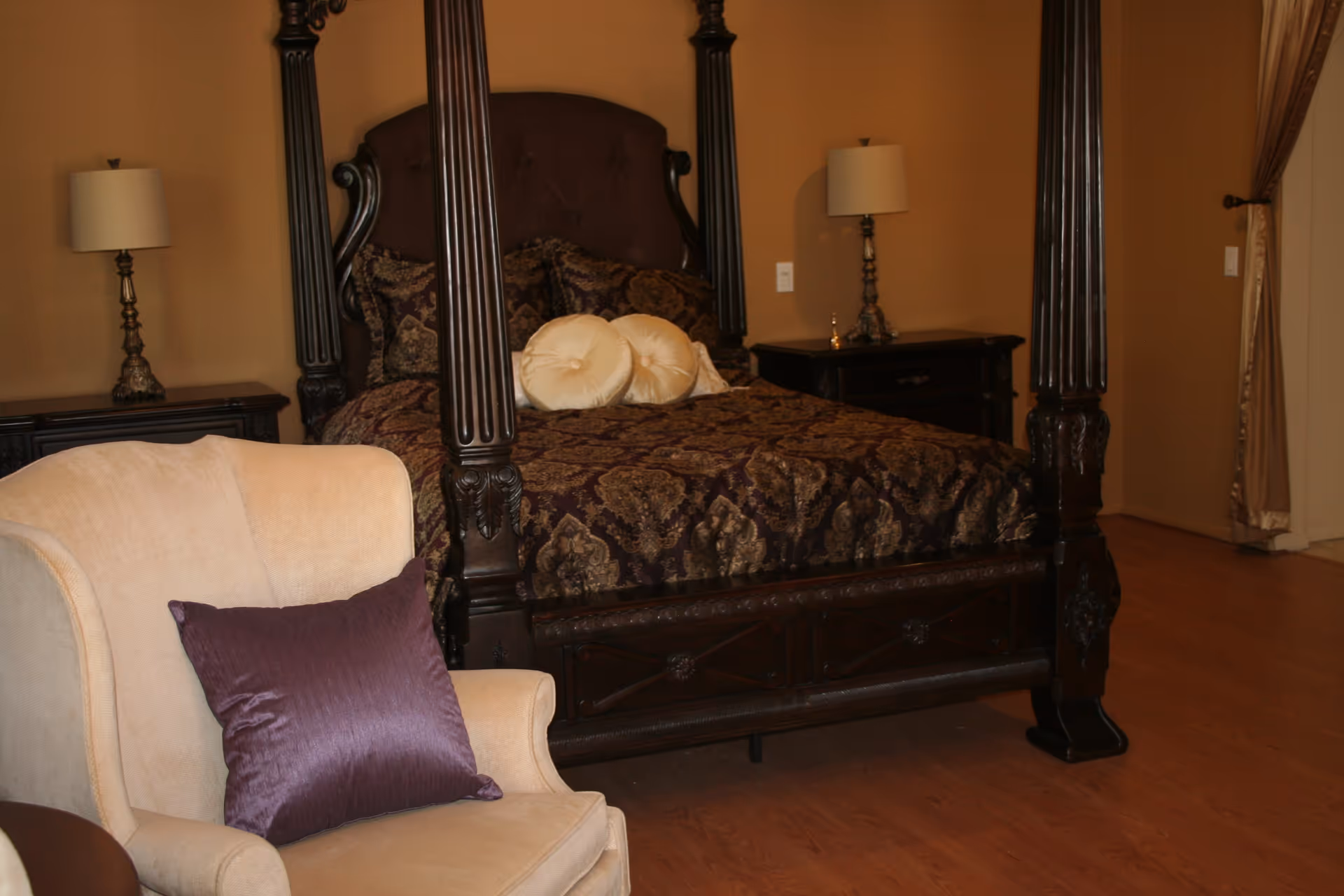 A bedroom with a dark wooden four-poster bed adorned with patterned bedding and two round cream pillows. There are two matching bedside tables with lamps on each side of the bed. In the foreground, there is a beige armchair with a purple cushion. The room has wooden flooring and beige walls with a curtain partially visible on the right side.
