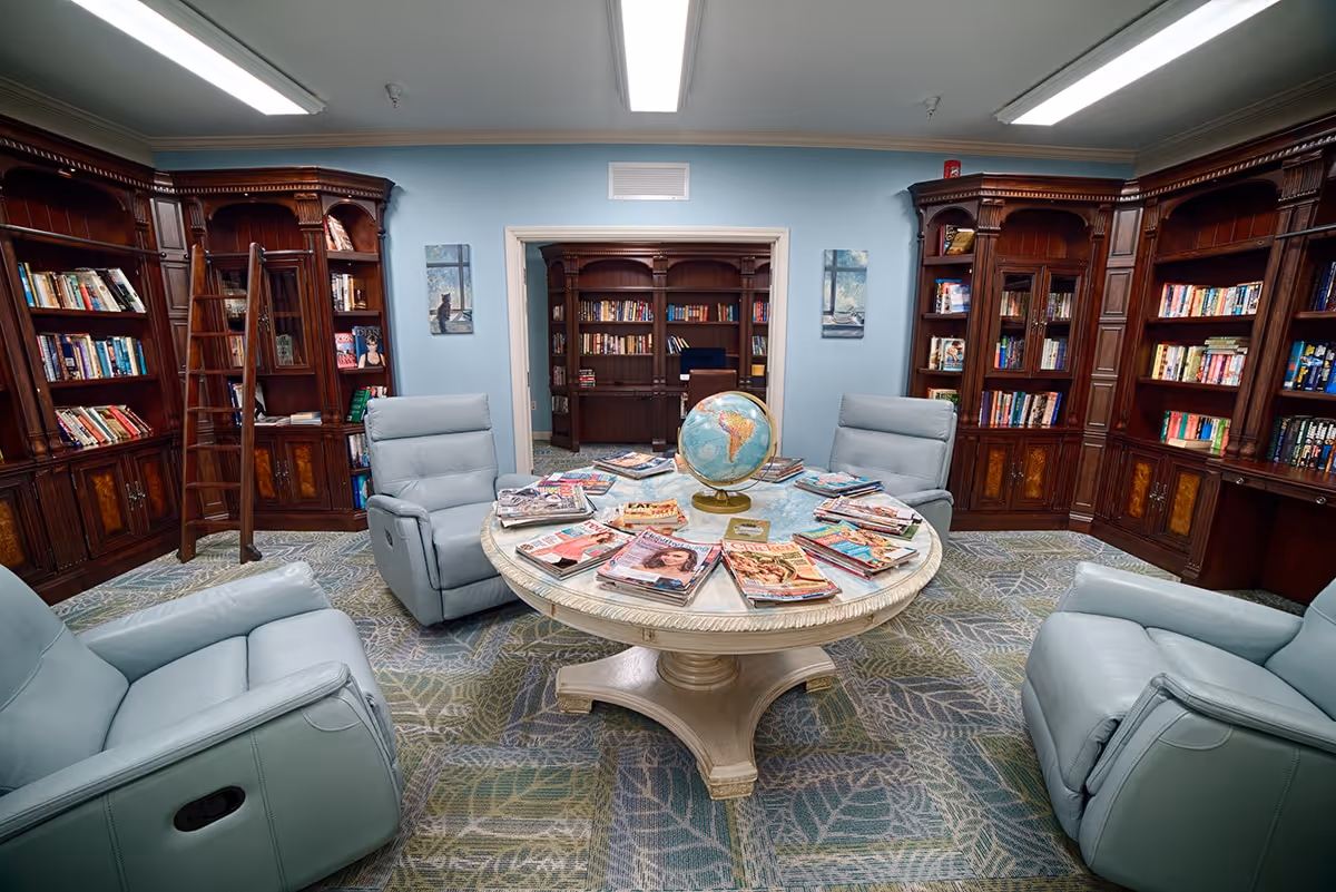 A cozy library room with light blue walls and carpet featuring a leaf pattern. The room contains four light blue recliner chairs arranged around a round wooden table with a globe and several magazines on it. The walls are lined with dark wooden bookshelves filled with books, and a rolling ladder is attached to one of the shelves. Through a doorway, another room with more bookshelves and a desk is visible.