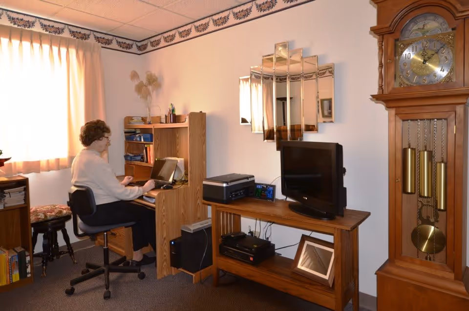 A woman sitting on an office chair working on a laptop at a wooden desk in a cozy room. The room has a window with curtains, a wooden grandfather clock, a TV on a wooden stand, a printer, and a decorative mirror on the wall.