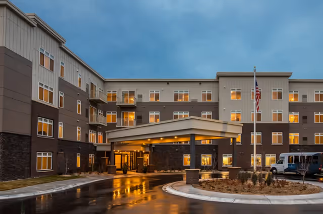 Exterior view of a modern senior living facility building at dusk with multiple windows lit from inside, a covered entrance with lights, a flagpole with an American flag, and a parked van near the entrance.