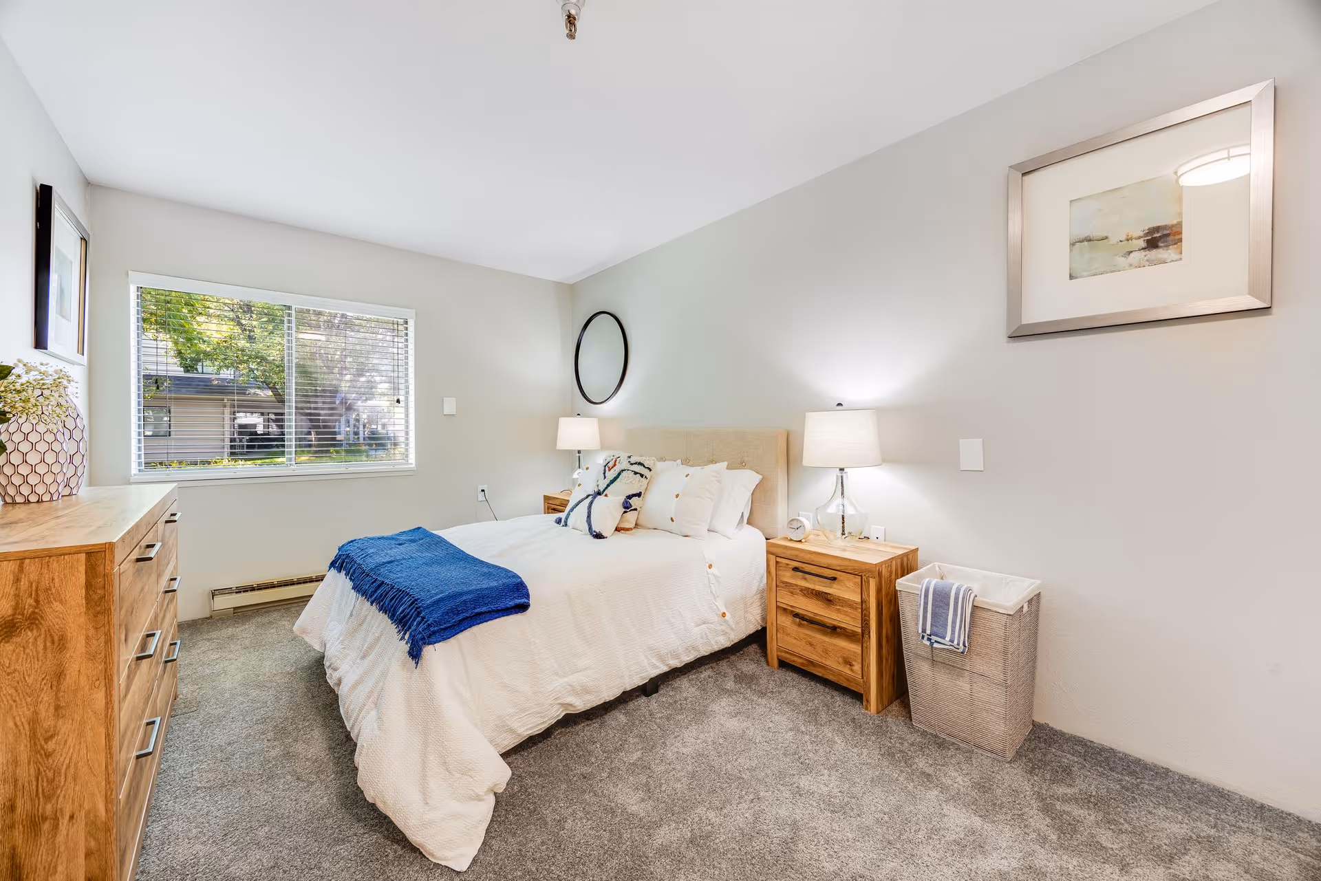 A bright and cozy bedroom with a large window letting in natural light. The room features a bed with white bedding and a blue throw blanket, two wooden nightstands with lamps, a wooden dresser, a round mirror on the wall, a framed picture, and a laundry basket with a towel draped over it. The carpet is gray and the walls are painted light gray.