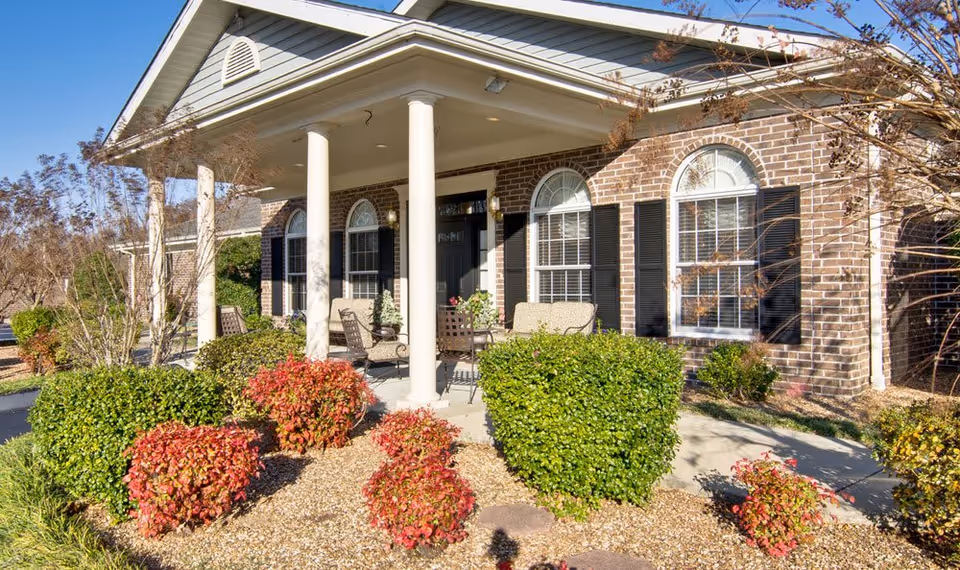 Exterior view of a brick building with a covered porch supported by white columns. The porch has outdoor seating with chairs and a small table. The surrounding landscaping includes green and red bushes, small trees, and a gravel ground cover under a clear blue sky.