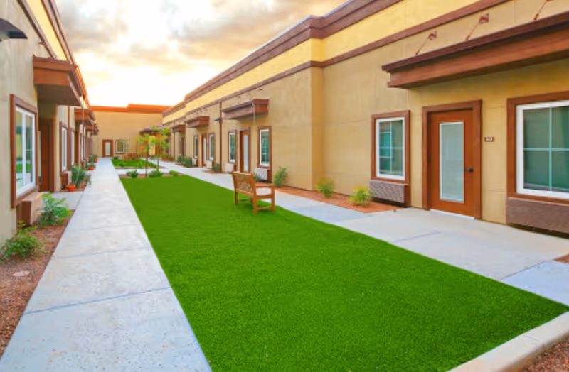 Outdoor courtyard area at Vineyard Park of Surprise featuring a green artificial grass lawn in the center, surrounded by a concrete walkway and beige building walls with windows and doors. There is a wooden bench on the grass and small plants along the walkway.