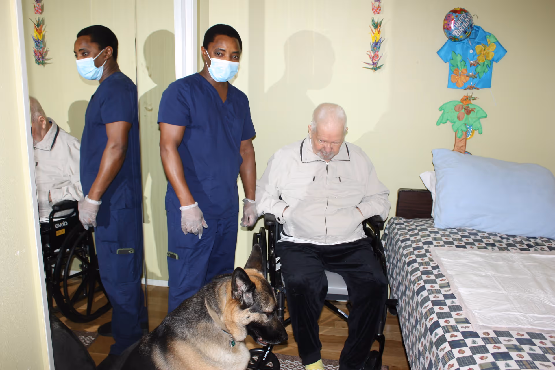 A healthcare worker wearing blue scrubs, a face mask, and gloves stands next to an elderly man in a wheelchair inside a room. A large dog, possibly a German Shepherd, is sitting in front of them. The room has a bed with a patterned bedspread and a pillow, and colorful decorations on the wall including a 'Happy Birthday' balloon.