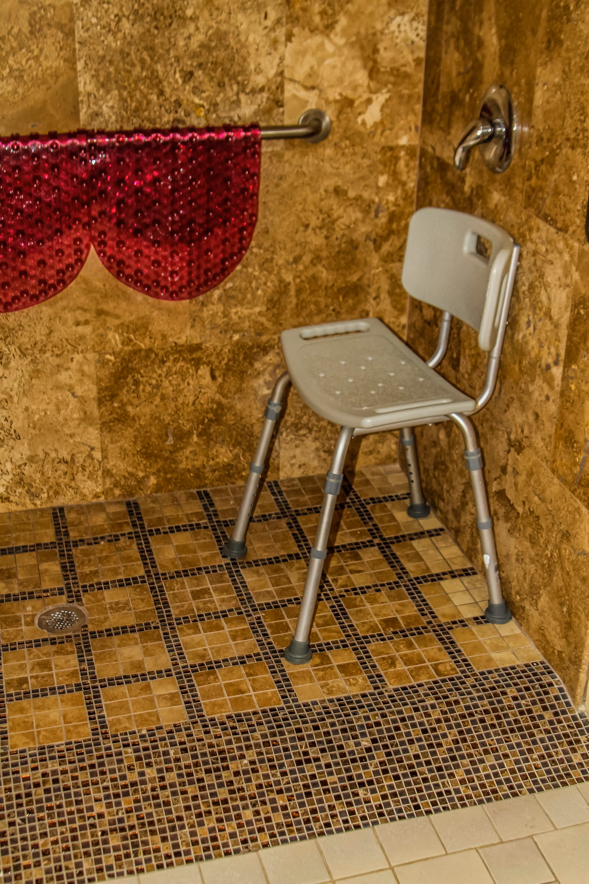 A shower area with brown tiled walls and floor, featuring a gray shower chair with backrest and a red textured towel hanging on a metal towel bar.