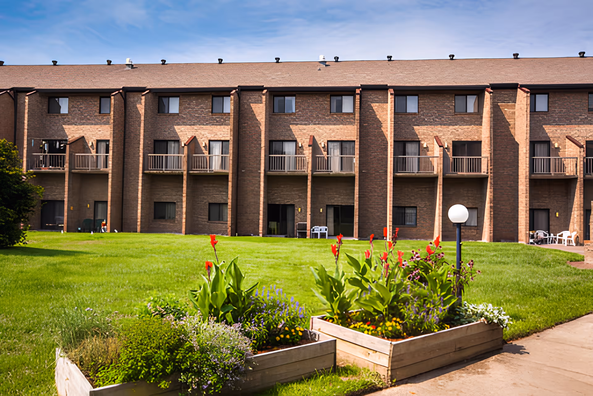 Exterior view of a two-story brick retirement community building with balconies and sliding glass doors. In the foreground, there is a well-maintained green lawn with raised garden beds containing various plants and flowers, and a round white lamp post.