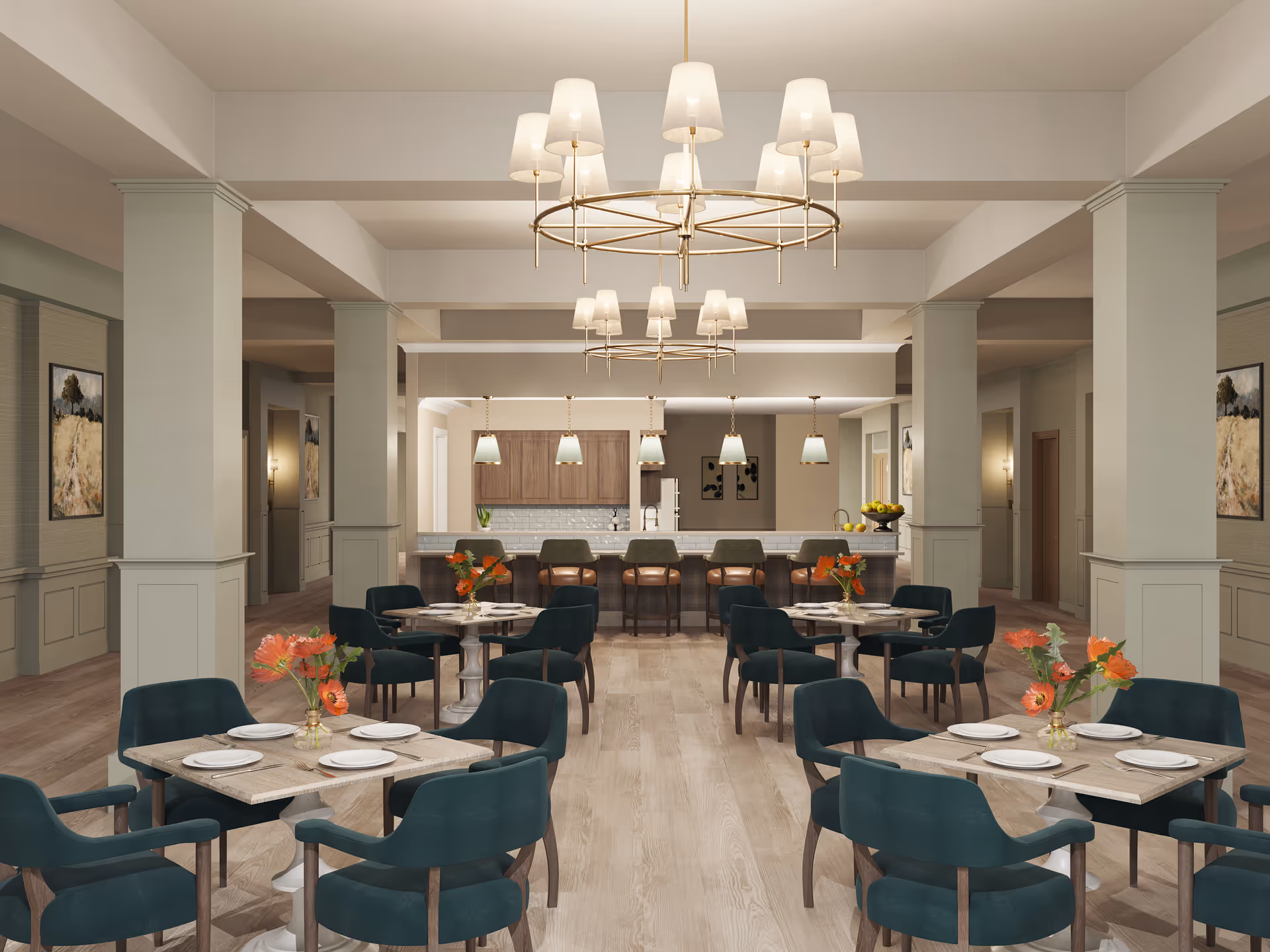A spacious dining area with multiple square tables set with white plates and orange flower centerpieces. Each table is surrounded by dark green upholstered chairs. In the background, there is a kitchen area with a long counter and five bar stools. The room features light wood flooring, beige walls with wainscoting, and two large circular chandeliers hanging from the ceiling. Artwork is displayed on the walls, and pendant lights hang above the kitchen counter.