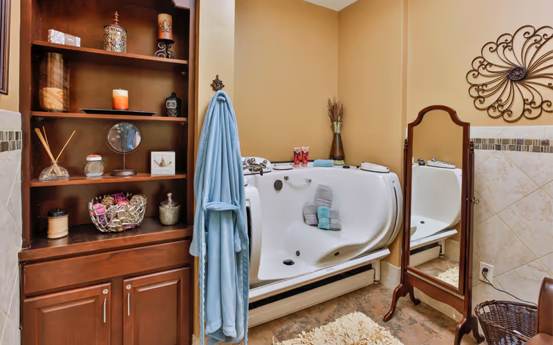 A cozy bathroom featuring a white walk-in bathtub with folded towels on the seat. To the left is a wooden shelving unit with decorative items and a blue bathrobe hanging on a hook. On the right side, there is a full-length wooden framed mirror and a decorative wall piece above beige tiled walls. The floor has a soft rug and a wicker basket is partially visible.