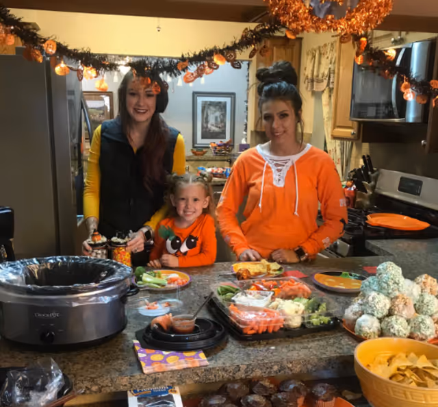 Three people, two women and a young girl, standing behind a kitchen counter decorated with Halloween-themed garlands. The counter is filled with various snacks including vegetables with dip, cupcakes, and a bowl of chips. The kitchen has wooden cabinets and stainless steel appliances.