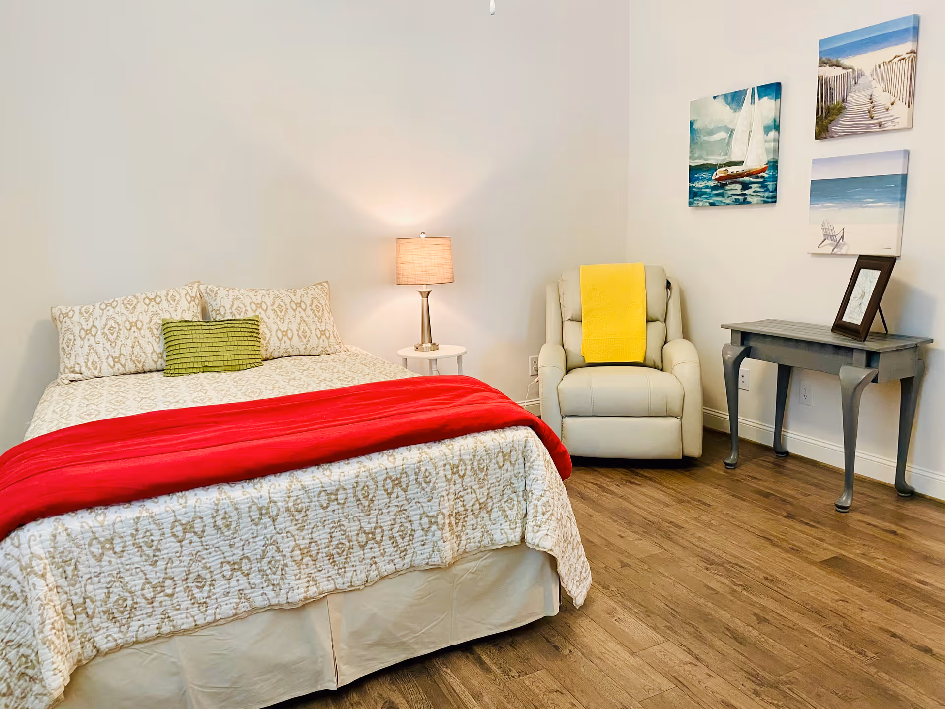 A cozy bedroom with a bed covered in a patterned beige and white bedspread, a red blanket, and a green pillow. Next to the bed is a small round white side table with a lamp. There is a beige recliner chair with a yellow blanket draped over it, a gray side table with a framed picture, and three beach-themed paintings on the wall. The floor is wooden.