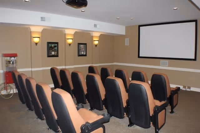 A small theater room with three rows of tan and black cushioned seats facing a large white projection screen mounted on a beige wall. The room has wall sconces providing soft lighting, framed pictures on the walls, and a popcorn machine in the corner.