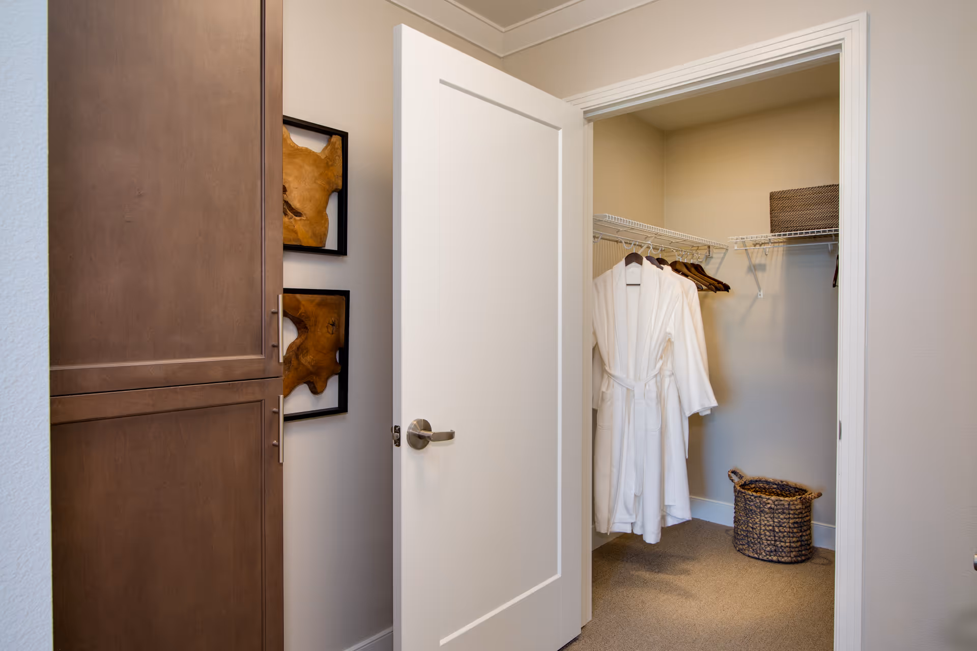 Open white door revealing a walk-in closet with white bathrobes hanging on wooden hangers, a woven basket on the carpeted floor, and wire shelving with a brown storage basket. To the left of the door is a tall wooden cabinet and two framed wooden art pieces on the wall.