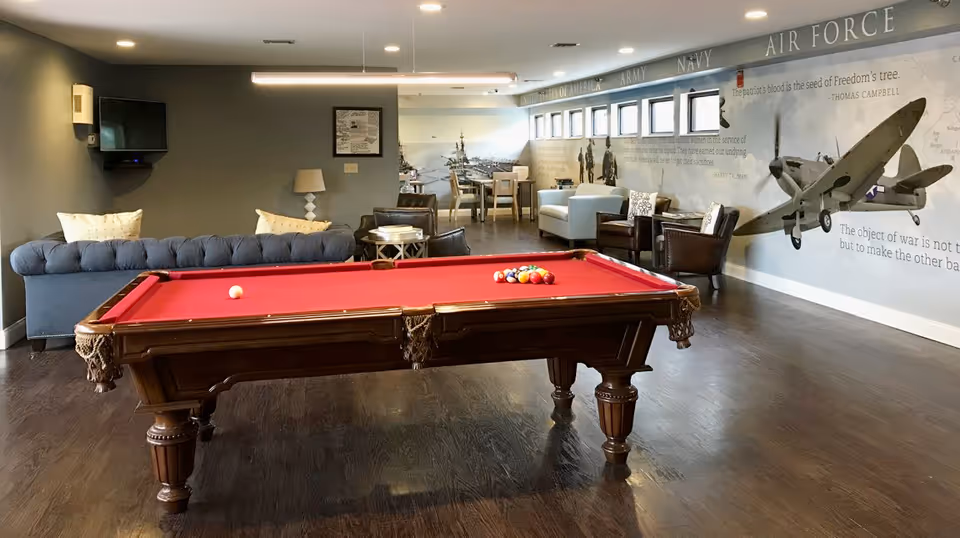 A spacious lounge area with a red felt pool table in the center. Behind the pool table, there is a gray tufted sofa with two pillows, a small side table with a lamp, and a wall-mounted TV. Further back, there are several chairs and tables arranged near a wall decorated with military-themed murals and quotes, including an airplane and references to the Army, Navy, and Air Force. The room has wooden flooring and multiple small windows along the mural wall.
