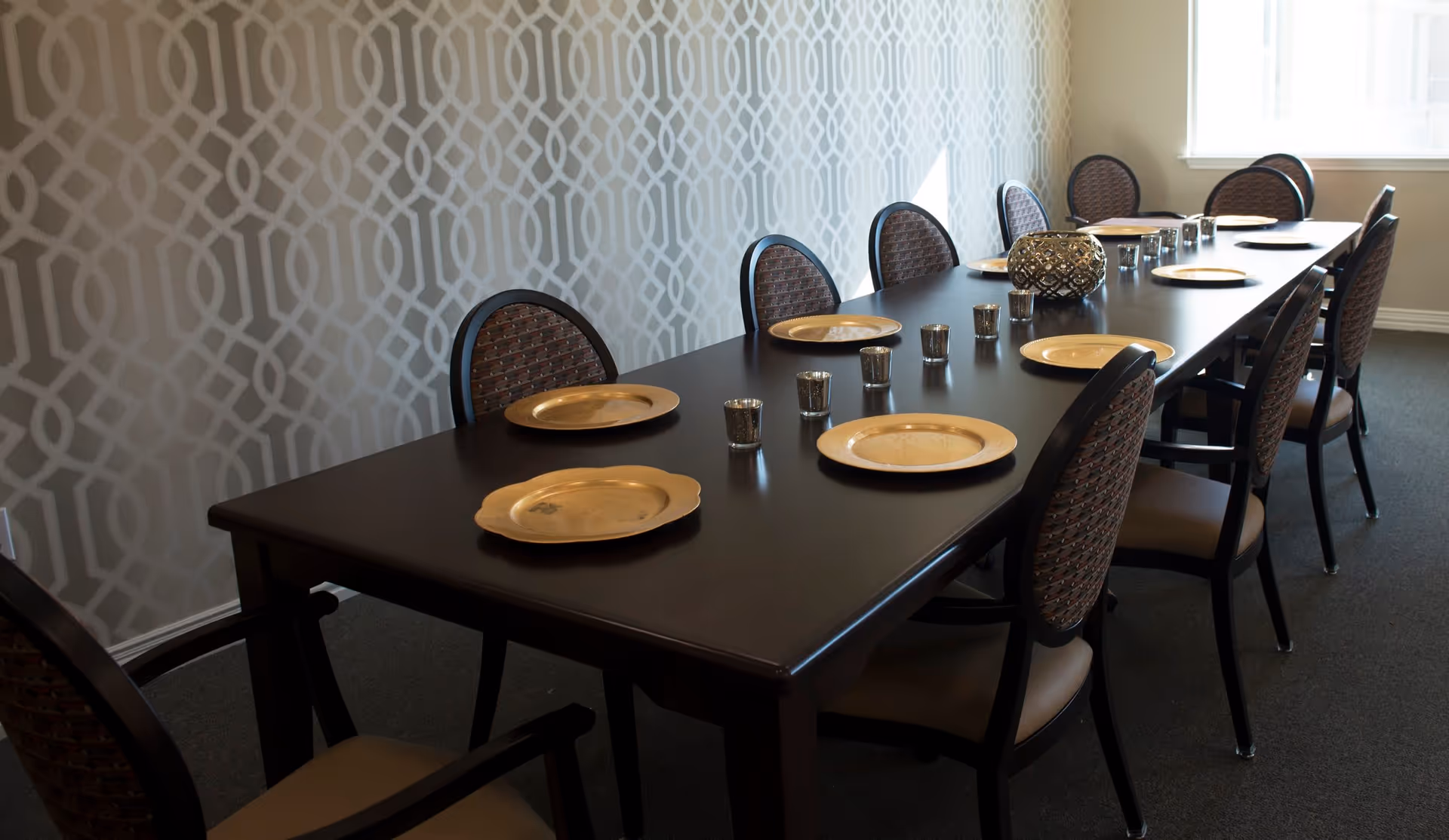 A long dark wooden dining table set with gold-colored plates and small candle holders, surrounded by chairs with patterned backs and cushioned seats, against a wall with a geometric patterned wallpaper and a window letting in natural light.