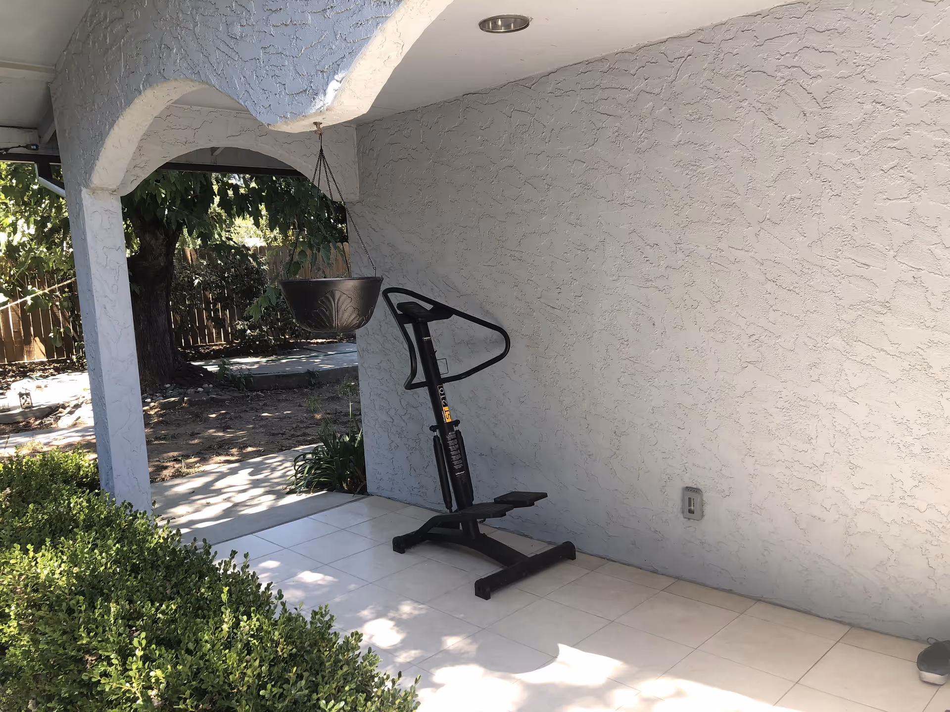 Outdoor covered patio area with a hanging planter and a black exercise stepper machine against a textured light gray wall. There are green bushes in the foreground and a tree with a wooden fence in the background.
