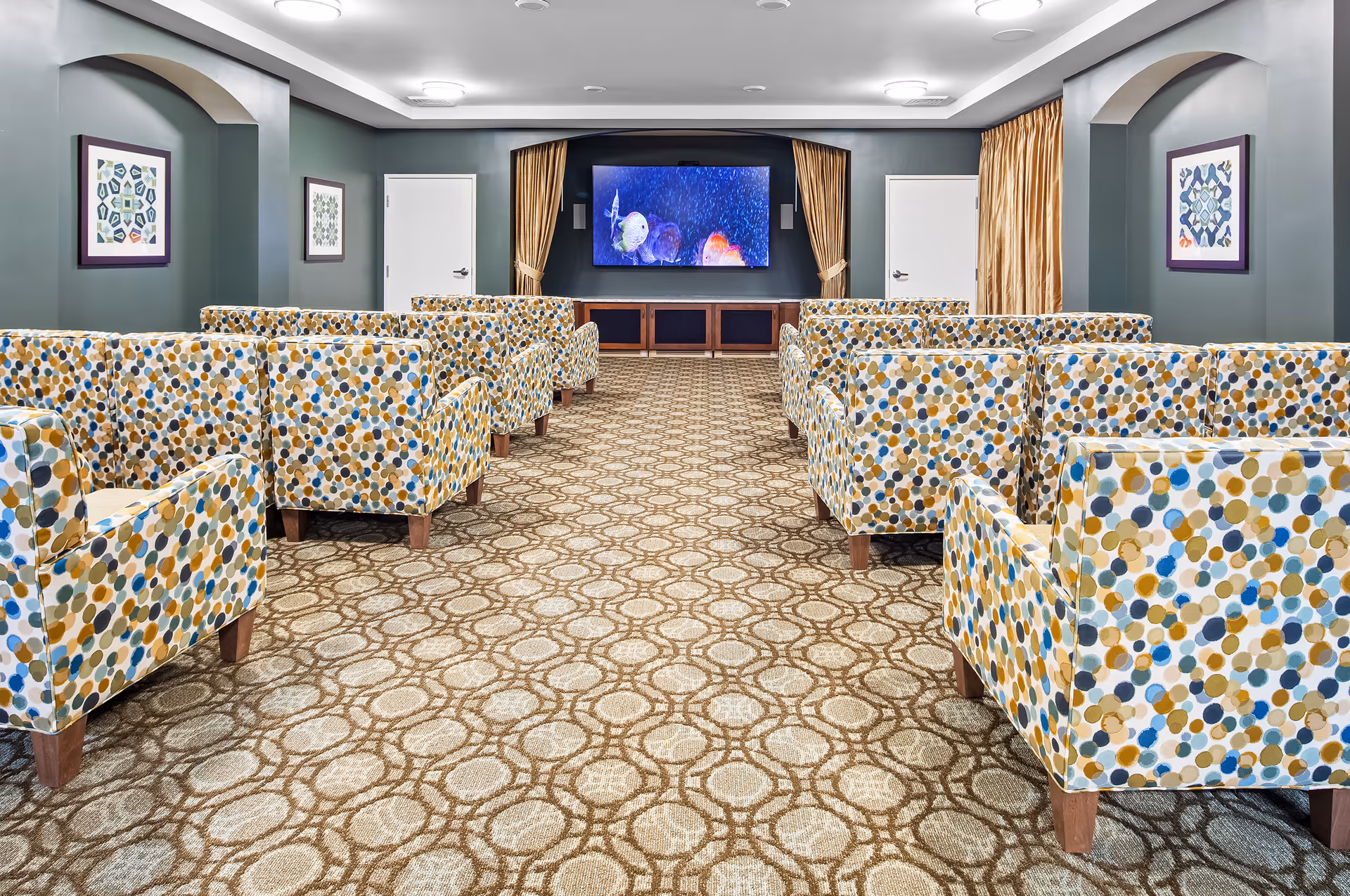 A cozy media room with patterned armchairs arranged in rows facing a large flat-screen TV mounted on a dark accent wall. The room features a patterned carpet, framed artwork on the walls, two white doors on either side of the TV, and gold curtains framing the TV area.