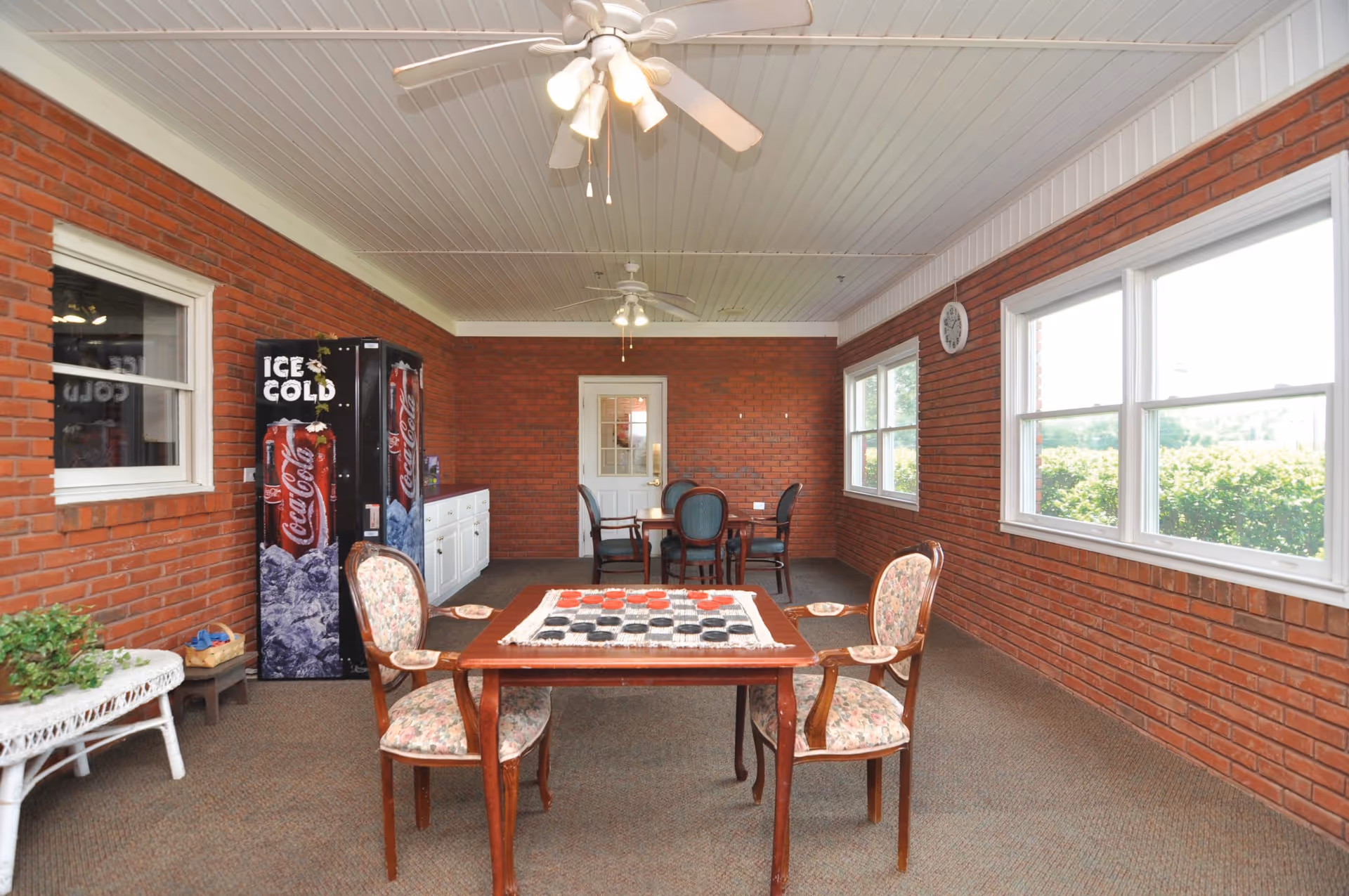 A bright room with red brick walls and large windows letting in natural light. The room features a wooden table with a checkerboard game set up and two floral upholstered chairs. In the background, there is another table with four chairs, a white cabinet, and a Coca-Cola vending machine. The ceiling has white paneling with two ceiling fans and lights.