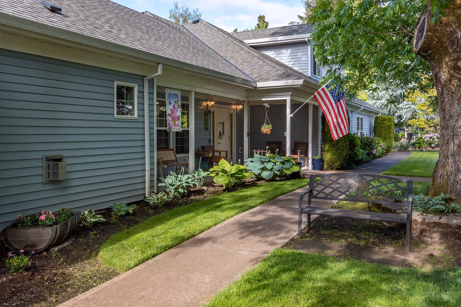 Exterior view of a single-story building with light gray siding, a sloped roof, and a small covered porch area. The porch has chairs and hanging plants, and an American flag is displayed on a pole attached to the building. A paved walkway runs alongside a well-maintained lawn and garden beds with various plants and flowers. A metal bench is positioned near a large tree in the foreground.
