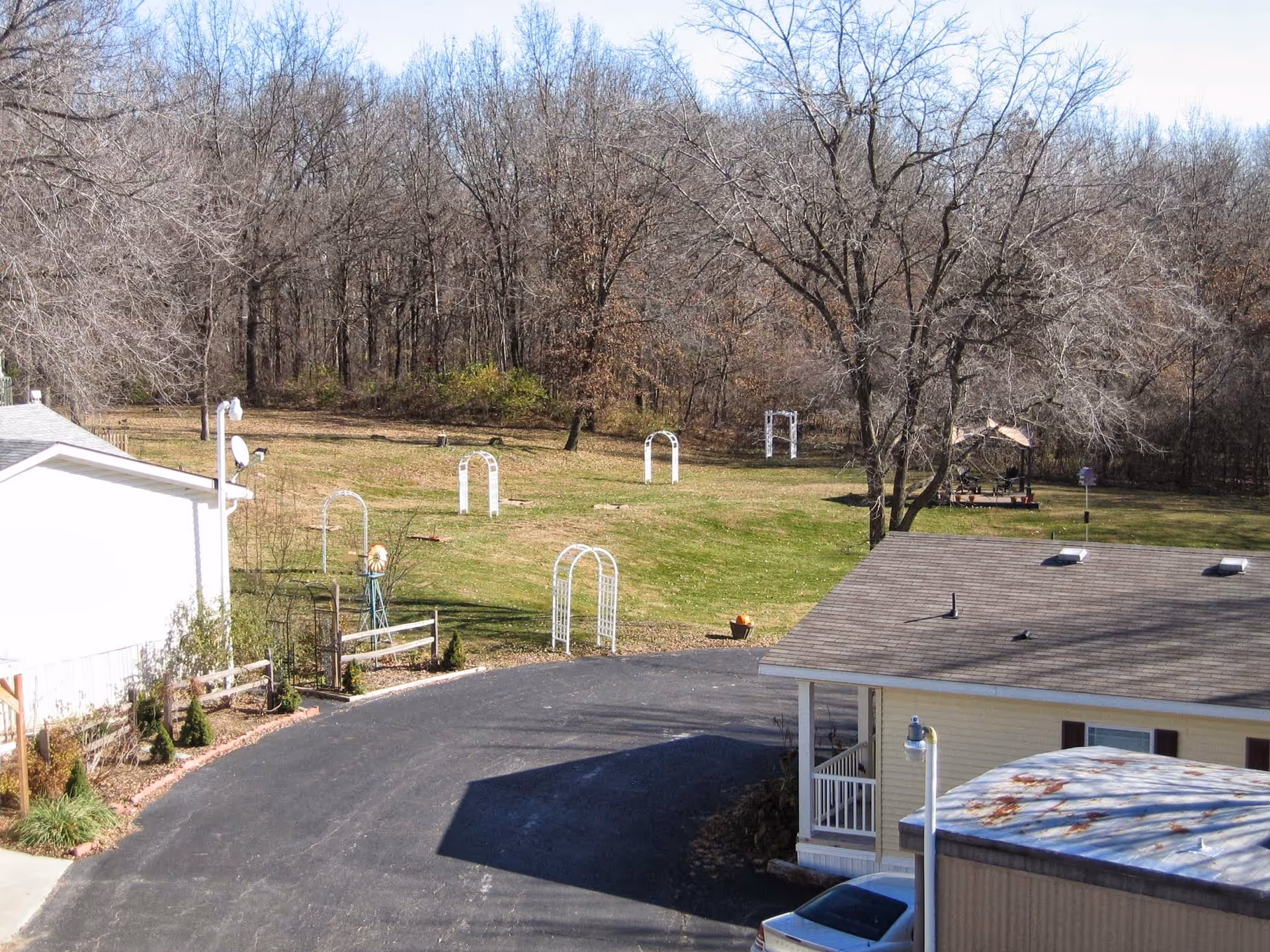 View of an outdoor area at Westwood Manor featuring a paved driveway, a grassy lawn with several white garden arches, leafless trees, and a small gazebo in the background. There are buildings on the left and right sides of the image.