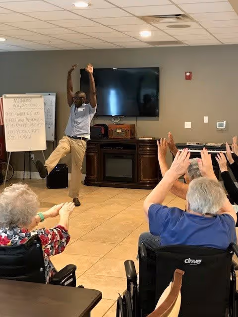 A man is leading a seated group of elderly people in an exercise or stretching activity in a community room. The man stands with one leg raised and arms extended upwards, while the elderly participants, some in wheelchairs, mimic his movements with their arms raised. The room has a large flat-screen TV mounted on the wall, a wooden cabinet, and a whiteboard with a schedule written on it.