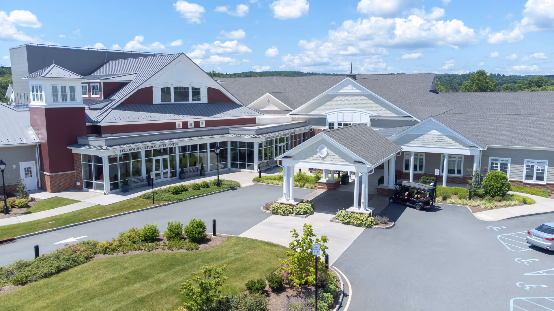Exterior view of Fellowship Village senior living facility showing a large building with a covered entrance, landscaped greenery, parking spaces, and a clear blue sky with some clouds.
