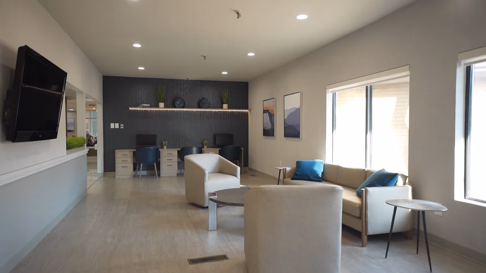 A modern and spacious common area with light-colored flooring and walls. The room features a beige sofa with blue cushions, two beige armchairs, and two small round tables. On the far wall, there are two desks with blue chairs and decorative plants and plates on a shelf above. Two framed artworks hang on the wall near large windows letting in natural light. A flat-screen TV is mounted on the left wall.