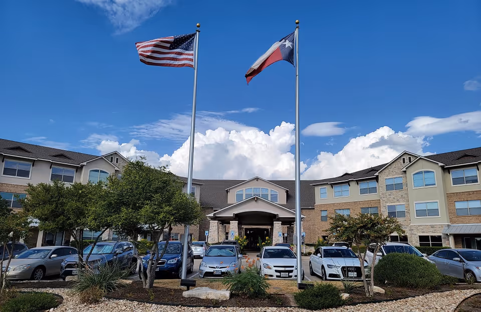 Front exterior of a multi-story senior living building with two flagpoles (American and Texas), parked cars, and landscaped grounds under a blue sky.