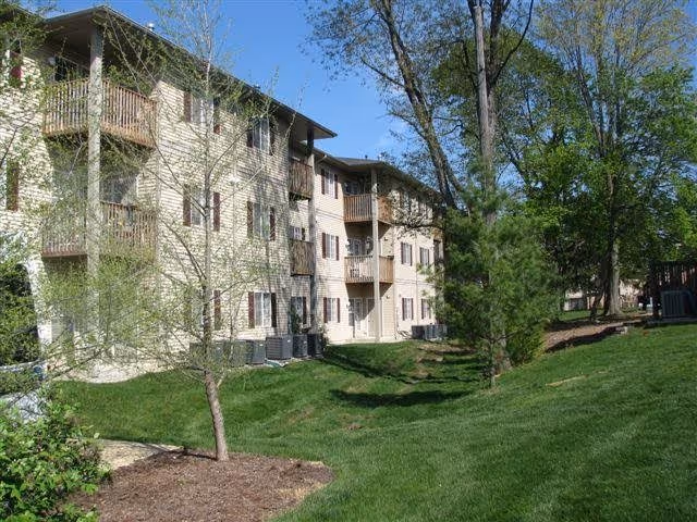 Exterior view of a multi-story residential building with balconies, surrounded by green grass, trees, and landscaping under a clear blue sky.