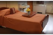 Senior living bedroom with a single bed covered by an orange bedspread, a tray with flowers on the bed, a wall-mounted sink, and a heater under the window.