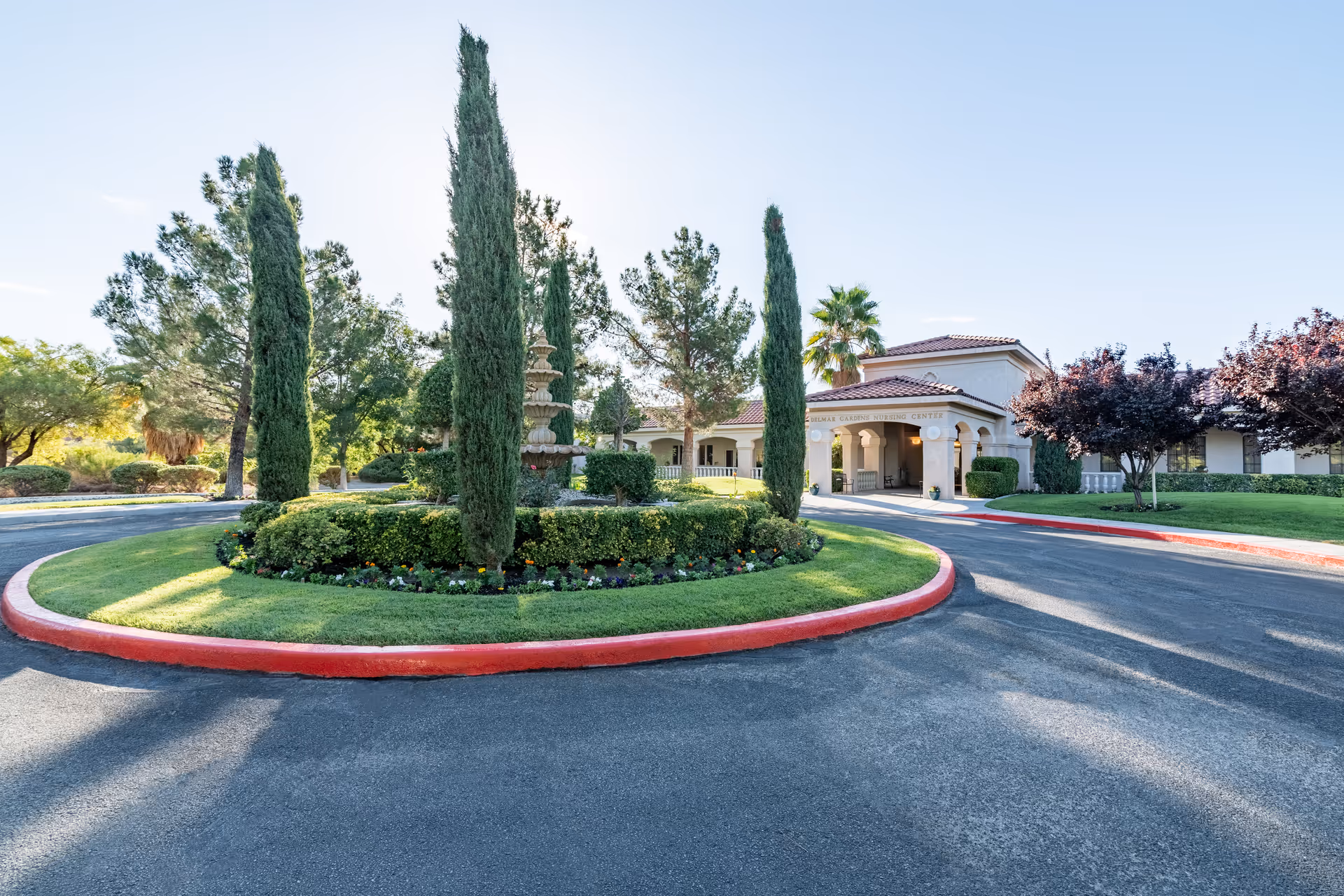 Building entrance with a circular driveway and landscaped roundabout featuring a fountain and tall cypress trees.
